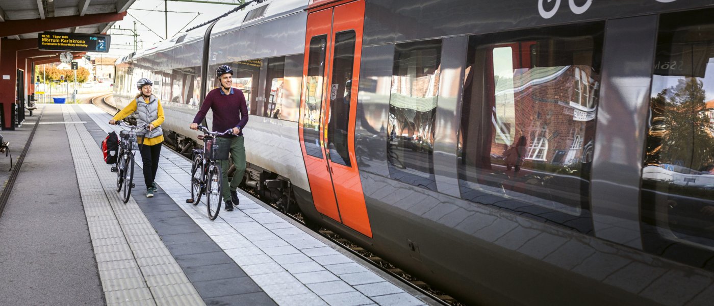Tågstation med ett tåg som står inne vid perrongen. Två personer leder sina cyklar bredvid. Foto.